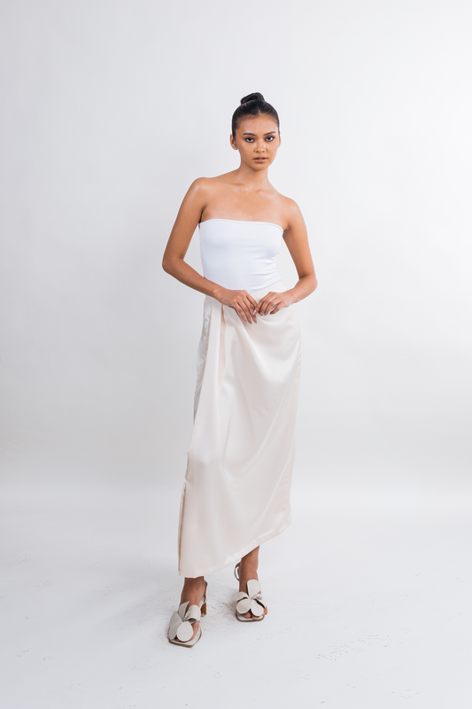 A woman modeling a strapless white dress with a gathered bodice and a flowing satin-like skirt, paired with white strappy sandals, against a seamless light gray studio background.