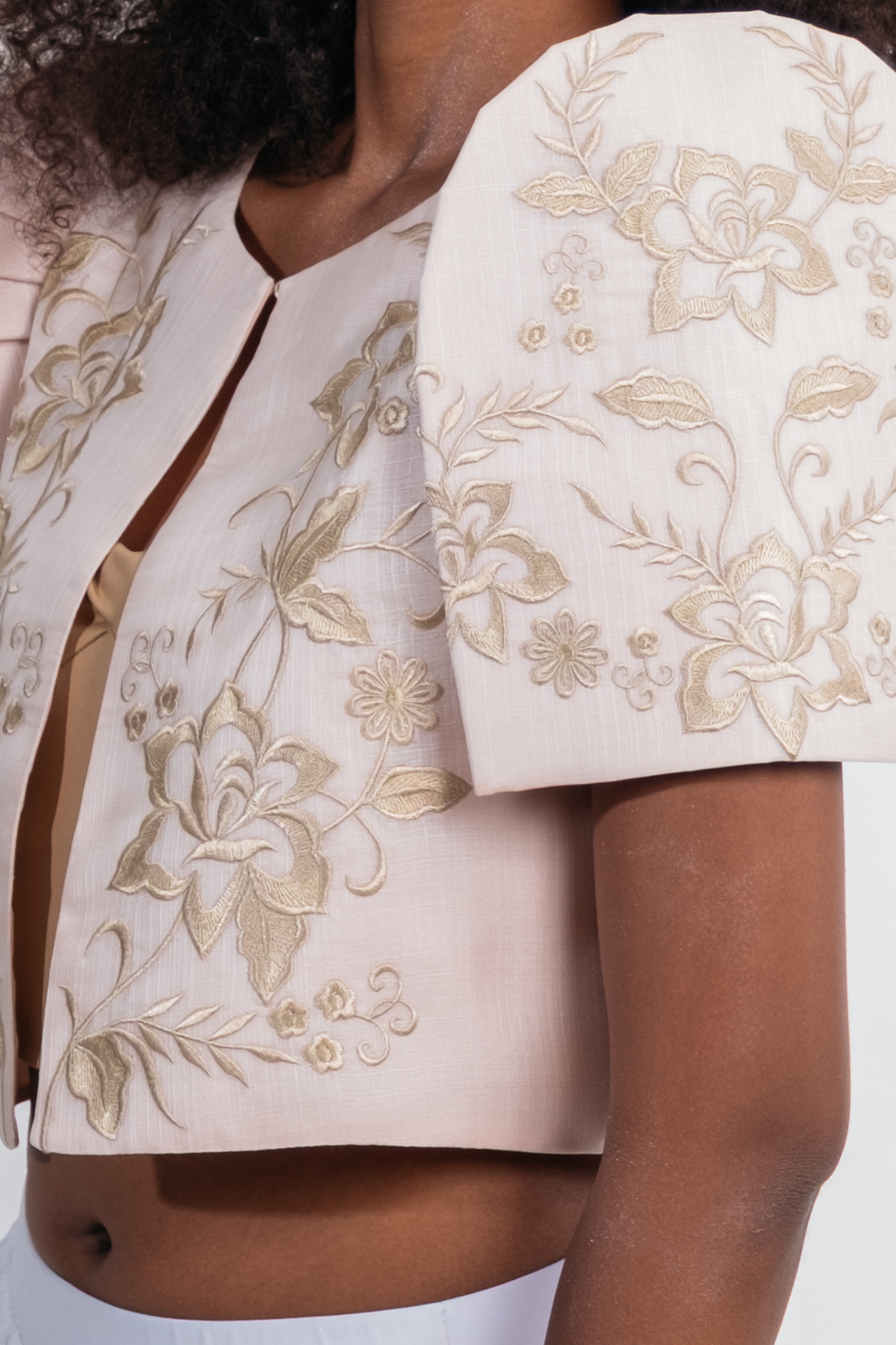Close-up of a model wearing a light beige bolero cropped jacket with gold floral embroidery and structured shoulder details.
