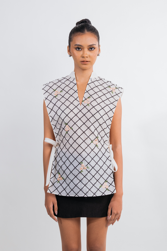 A model wearing a sleeveless white top with a black grid pattern and small floral accents, paired with a black skirt, against a light gray studio background.