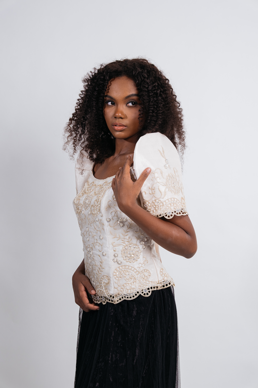 A woman with curly dark hair models a short-sleeve lace top in cream with a black skirt against a light gray studio background.