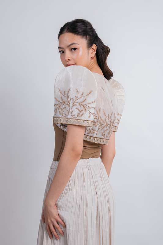 A woman wearing a light beige and brown traditional-style blouse with voluminous short butterfly sleeves and embroidered detailing, paired with a pleated light skirt, photographed against a plain light gray studio background.