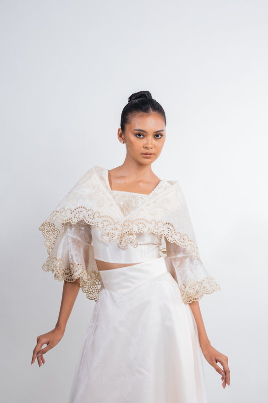A model wearing a white and cream lace and satin traditional-style outfit, featuring a high-neck crop top with dramatic ruffled sleeves and a matching skirt, set against a plain light gray studio background.