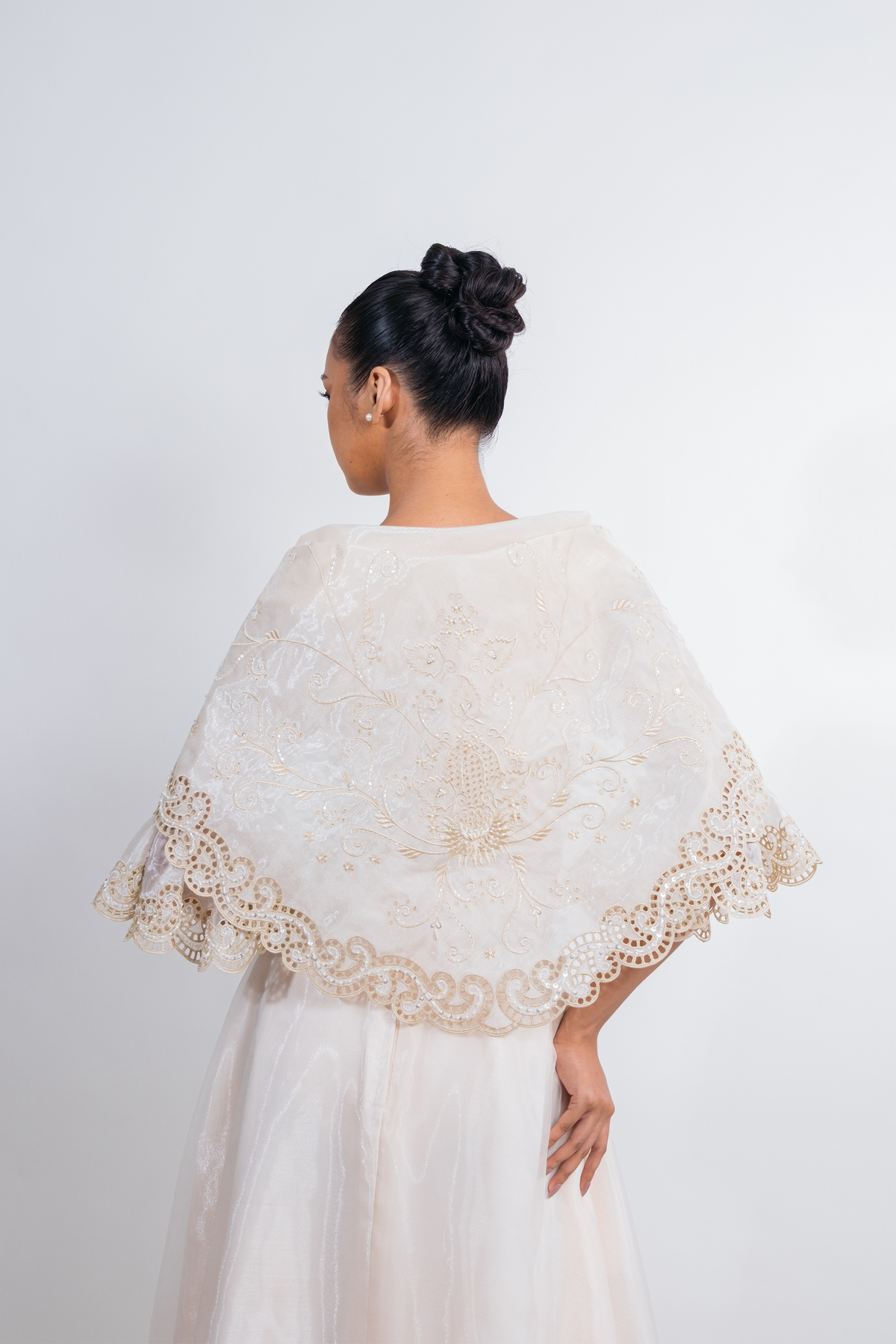 A woman shown from the back wearing a cream lace alampay or shawl over a white dress against a plain light gray studio background.