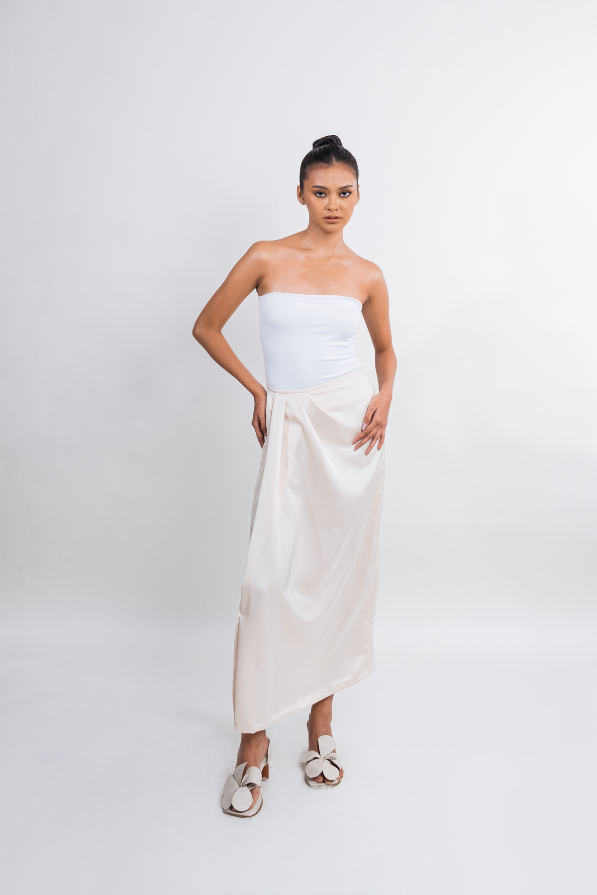 A model stands against a light gray studio background wearing a white strapless tube top and a long, draped off-white skirt, paired with white slide sandals.