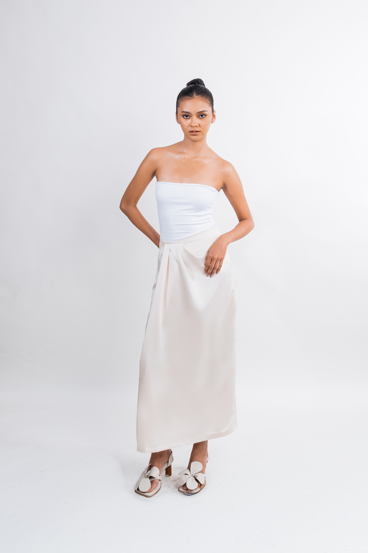 A woman modeling a strapless white tube top and a long, flowing cream or off-white skirt against a plain light gray studio background.