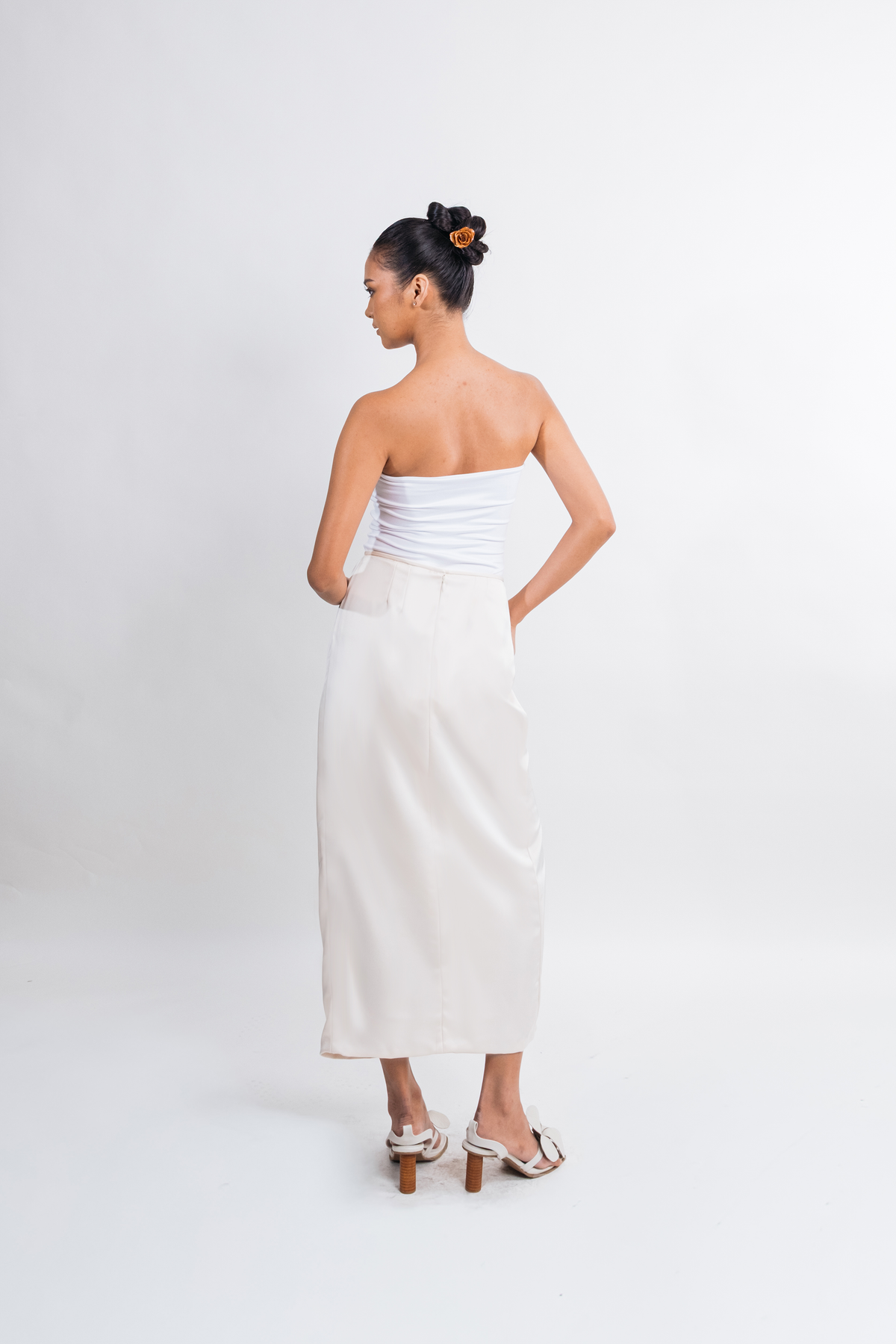A woman stands with her back to the camera wearing a strapless white tube top and a long white skirt, paired with white heeled sandals, against a plain light gray studio background.