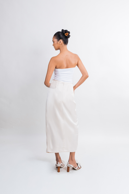 A woman stands with her back to the camera wearing a strapless white tube top and a long white skirt, paired with white heeled sandals, against a plain light gray studio background.