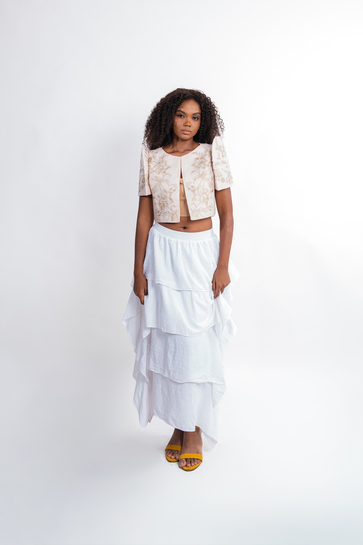 A model stands against a white studio background wearing a two-piece outfit consisting of a short-sleeve cropped top with a beige and white abstract pattern and a long white tiered skirt, paired with yellow sandals.