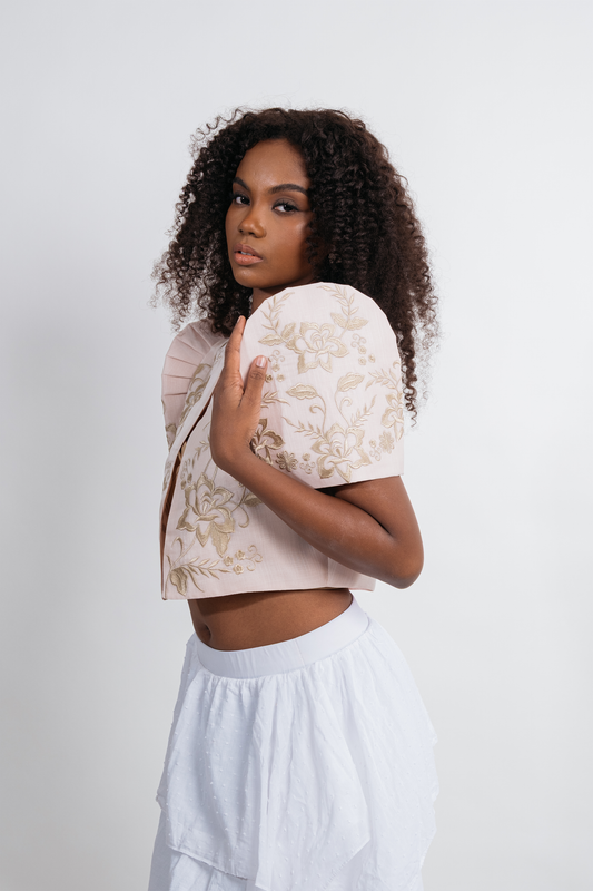 A model with curly dark hair is shown from the waist up, wearing a light beige butterfly sleeve bolero top with a gold floral pattern and a white skirt, set against a plain light gray studio background.