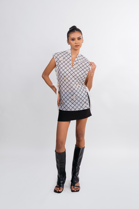 A model stands against a light gray studio background wearing a sleeveless wrap-style top with a black and white geometric grid pattern, a black mini skirt, and tall black heeled sandals.