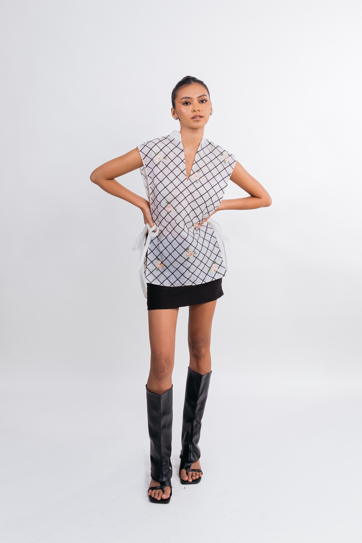 A model stands against a light gray studio background wearing a sleeveless wrap-style top with a black and white geometric grid pattern, a black mini skirt, and tall black heeled sandals.