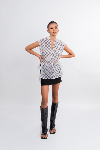 A model stands against a light gray studio background wearing a sleeveless wrap-style top with a black and white geometric grid pattern, a black mini skirt, and tall black heeled sandals.