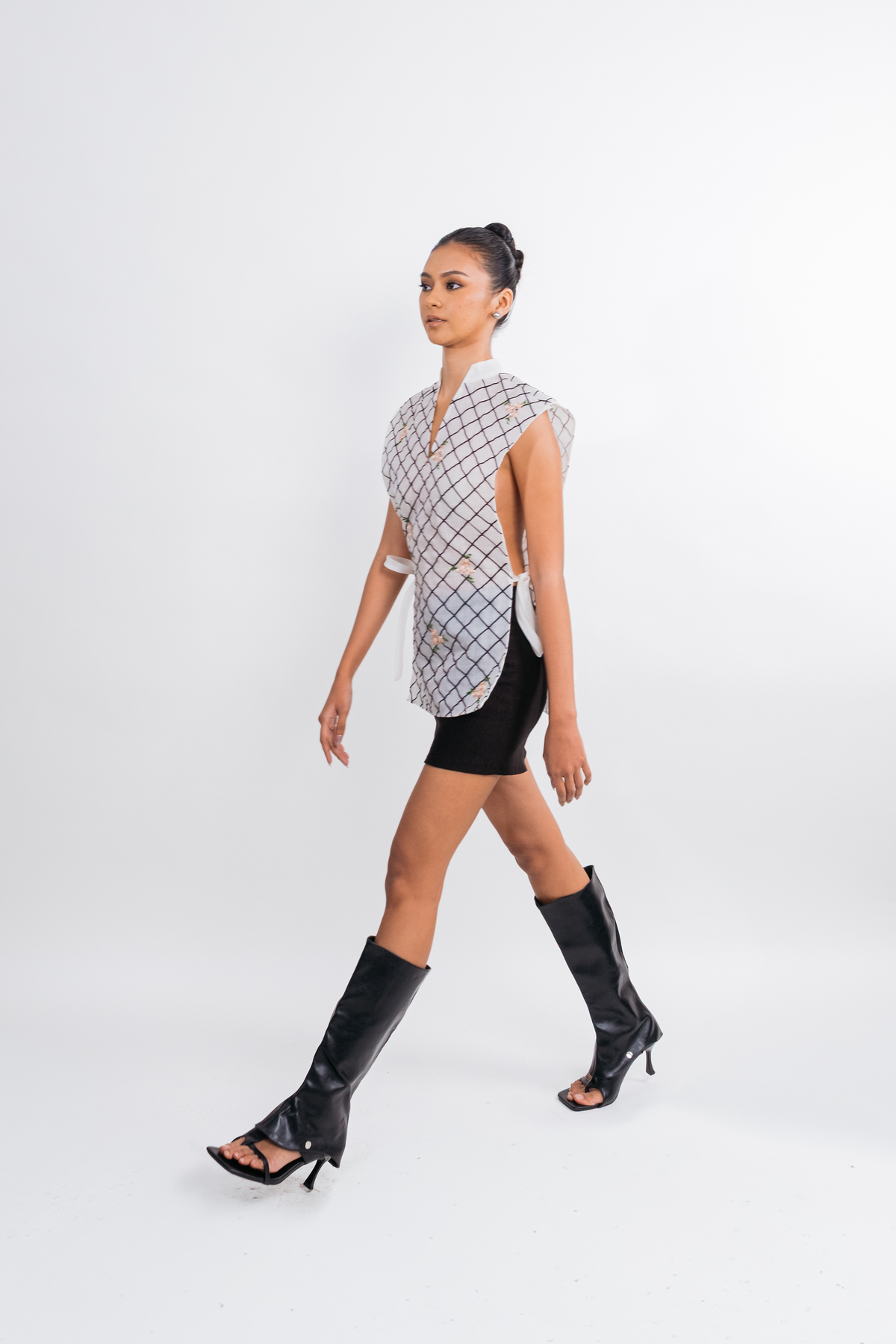 A model walks across a white studio background wearing a sleeveless black and white checkered vest, a black mini skirt, and black knee-high heeled sandals.