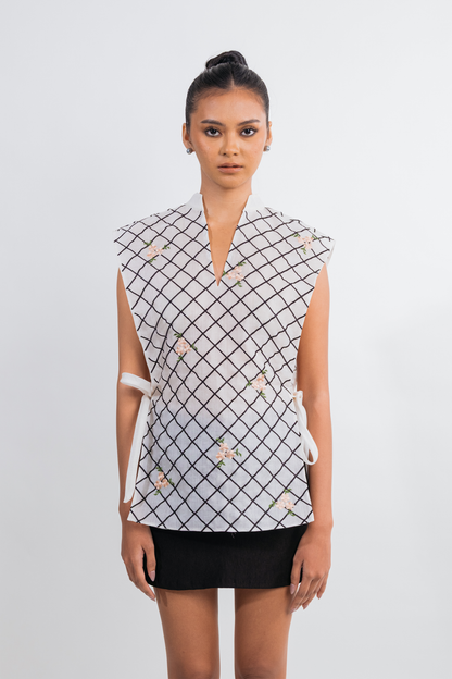 A model wearing a sleeveless white top with a black grid pattern and small floral accents, paired with a black skirt, against a light gray studio background.
