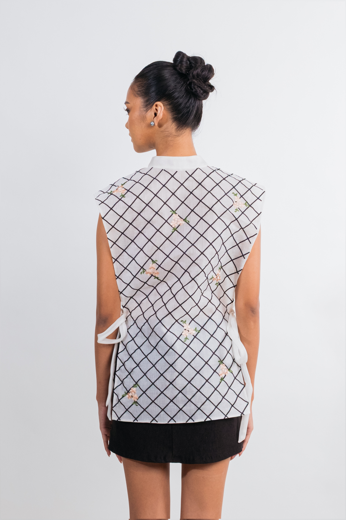 A woman shown from the back wearing a sleeveless white top with a black grid pattern and small pink floral accents, paired with a black skirt, against a light gray studio background.