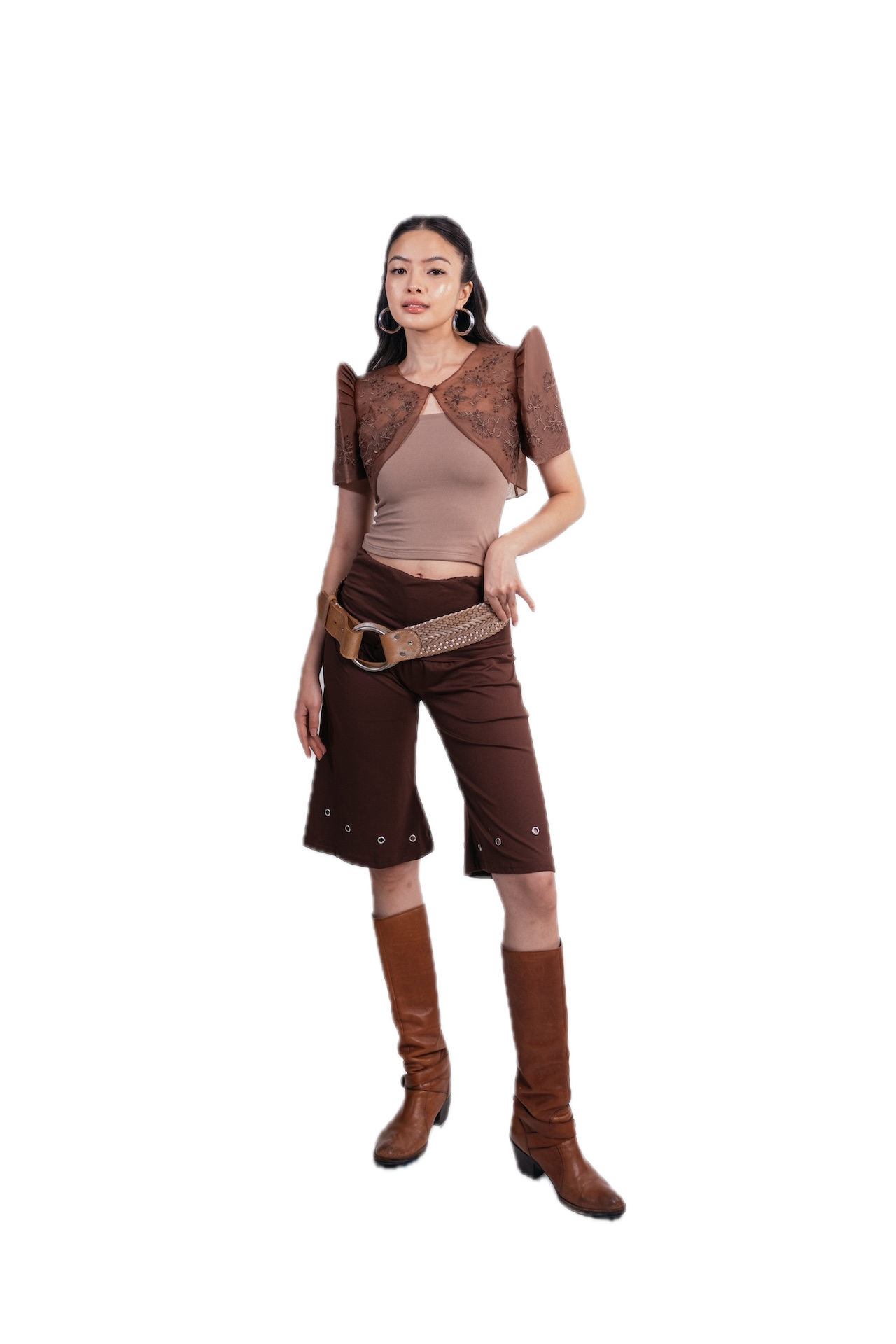 A woman modeling a brown and beige western-inspired outfit consisting of a patterned short-sleeve crop top, a beige ribbed tank top, brown cropped pants with silver studs, a woven belt, and brown knee-high boots, set against a plain white background.