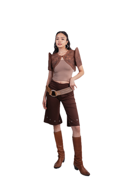 A woman modeling a brown and beige western-inspired outfit consisting of a patterned short-sleeve crop top, a beige ribbed tank top, brown cropped pants with silver studs, a woven belt, and brown knee-high boots, set against a plain white background.