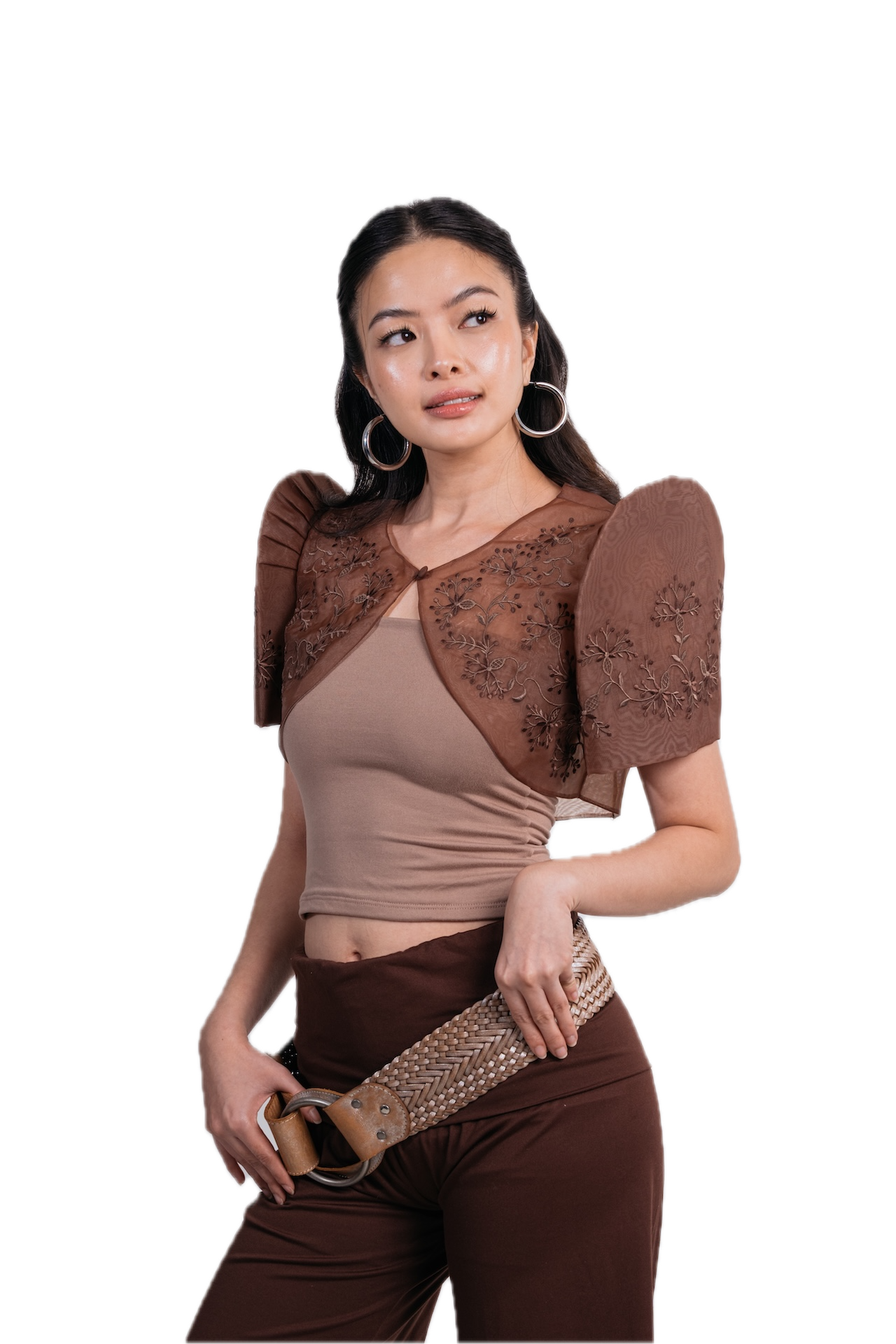 A woman modeling a brown sheer lace bolero shrug over a beige crop top and brown pants, holding a studded belt against a plain white background.