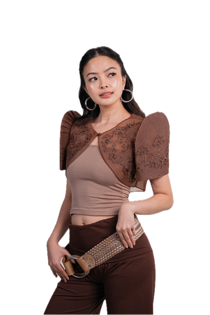 A woman modeling a brown sheer lace bolero shrug over a beige crop top and brown pants, holding a studded belt against a plain white background.