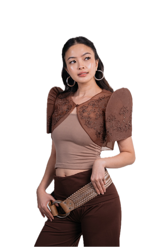 A woman modeling a brown sheer lace bolero shrug over a beige crop top and brown pants, holding a studded belt against a plain white background.
