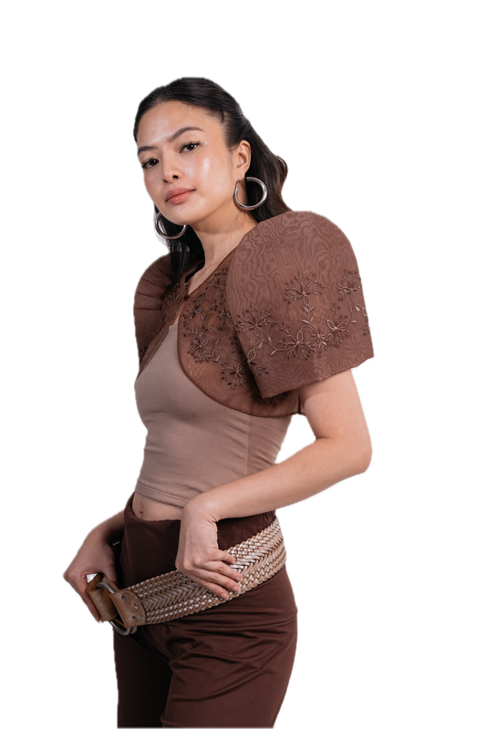 A woman modeling a brown sheer embroidered puff-sleeve shrug or cape over a beige top, paired with brown pants and a woven belt, against a solid white background.