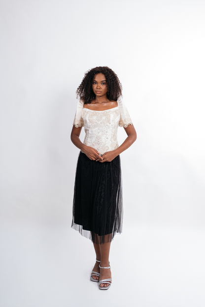 A woman stands against a plain light gray studio background wearing a short-sleeve off-the-shoulder white lace top and a long black tulle midi skirt with a sheer overlay, paired with silver heeled sandals.
