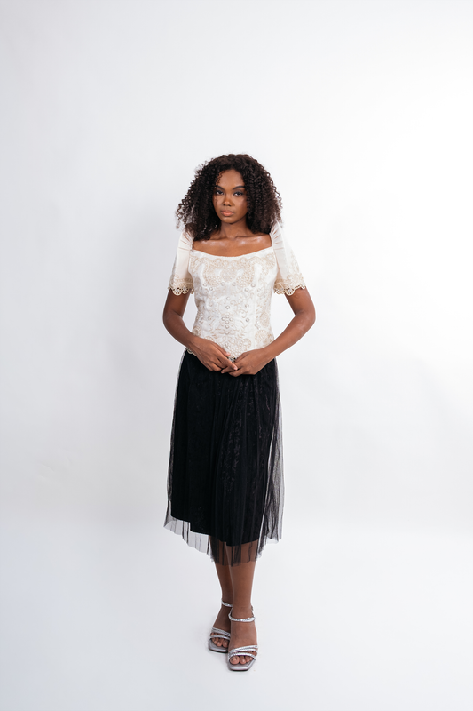 A woman stands against a plain light gray studio background wearing a short-sleeve off-the-shoulder white lace top and a long black tulle midi skirt with a sheer overlay, paired with silver heeled sandals.
