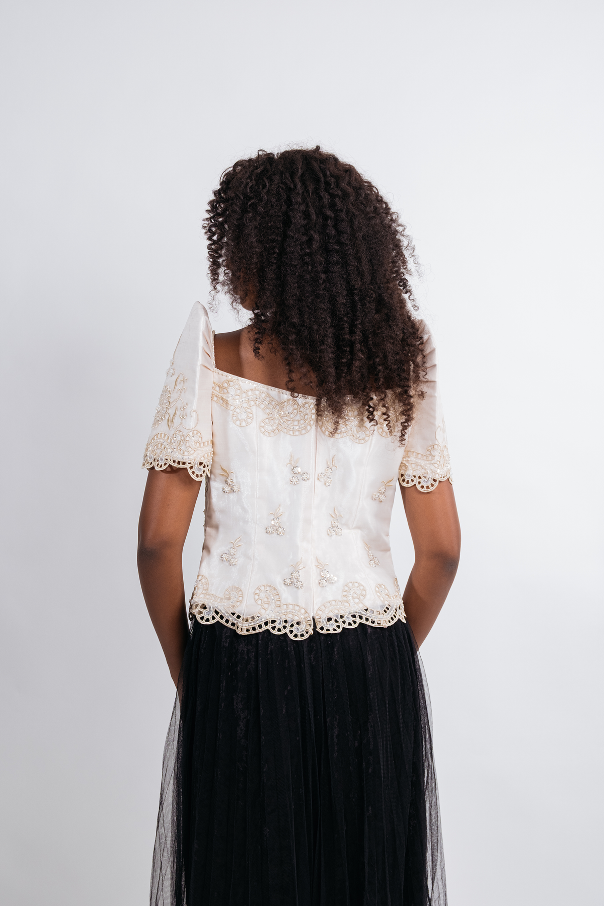 A woman viewed from behind wearing a cream lace short-sleeve top with a black tulle skirt, set against a plain light gray background.