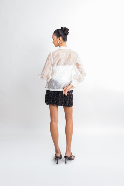A woman is shown from the back wearing a sheer white long-sleeve blouse, a black lace mini skirt, and black high heels against a plain white studio background.