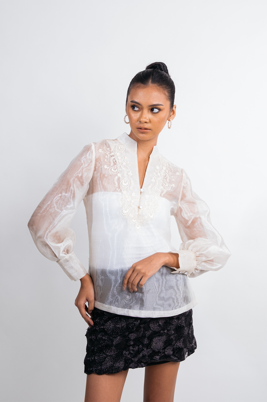 A woman modeling a white sheer lace blouse with long balloon sleeves and a black textured mini skirt, photographed against a plain light gray studio background.