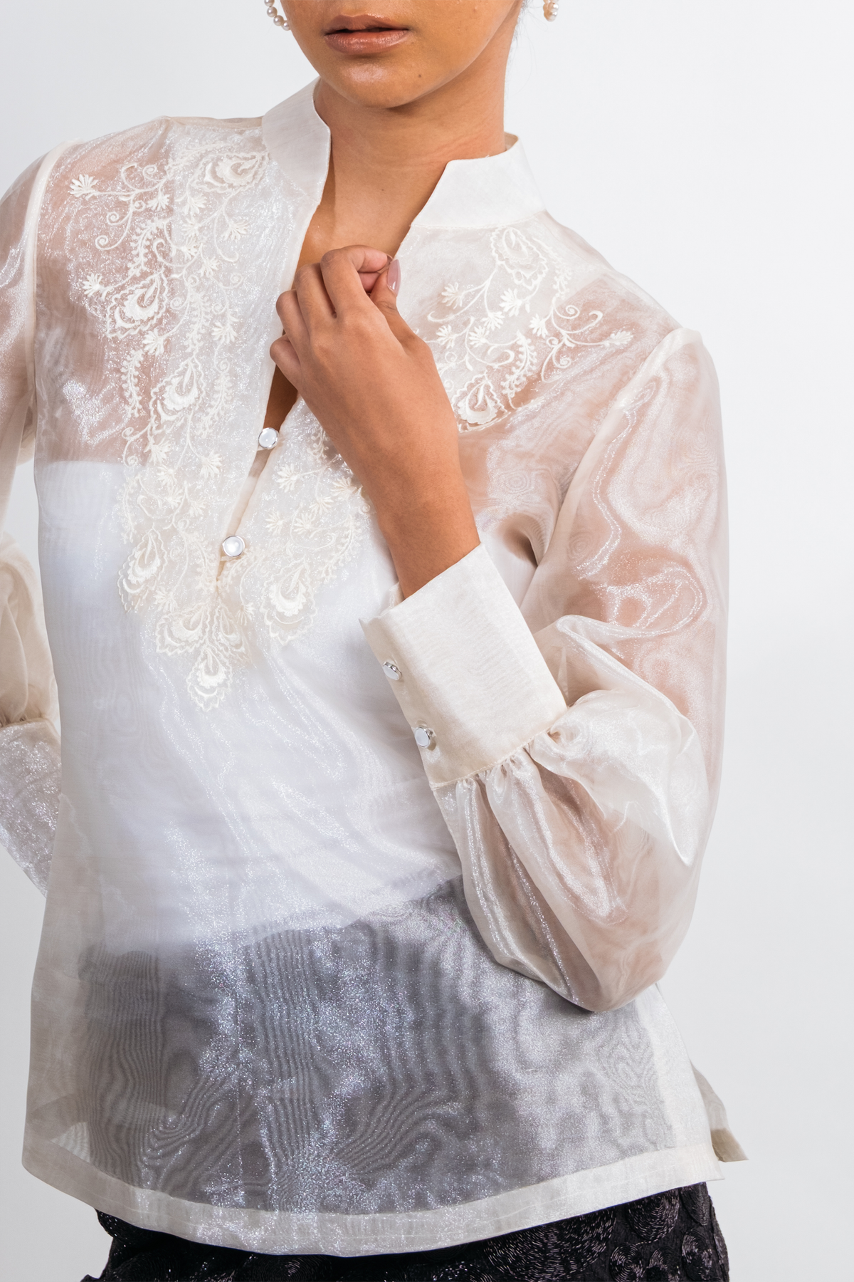 A close-up of a model wearing a white and silver embroidered blouse with sheer long sleeves and a high collar, set against a plain light background.