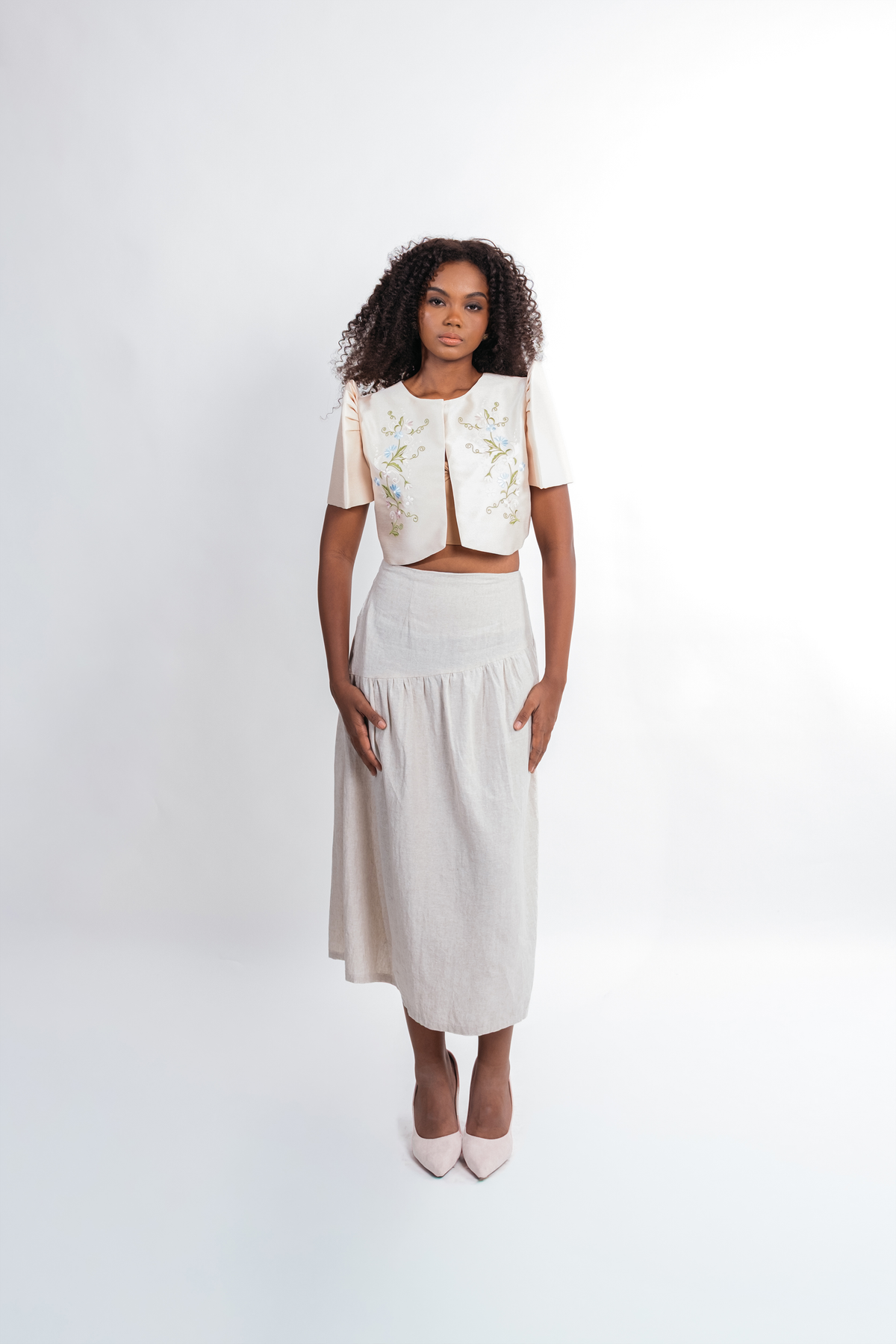 A model stands against a white studio background wearing a two-piece outfit consisting of a cream short-sleeve cropped bolero jacket with green floral embroidery and a matching cream midi skirt.