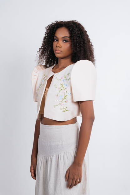 A model wearing a cream-colored cropped jacket with puff sleeves and delicate floral embroidery, paired with a matching cream skirt, against a plain light gray background.