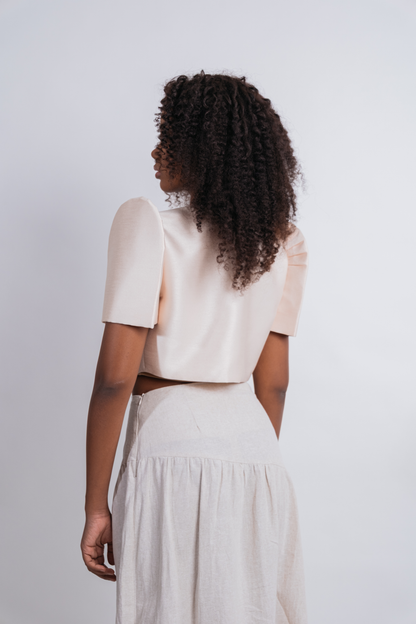 A woman with curly dark hair is shown from the back wearing a light cream short-sleeve crop top and a matching high-waisted linen skirt against a plain light gray background.