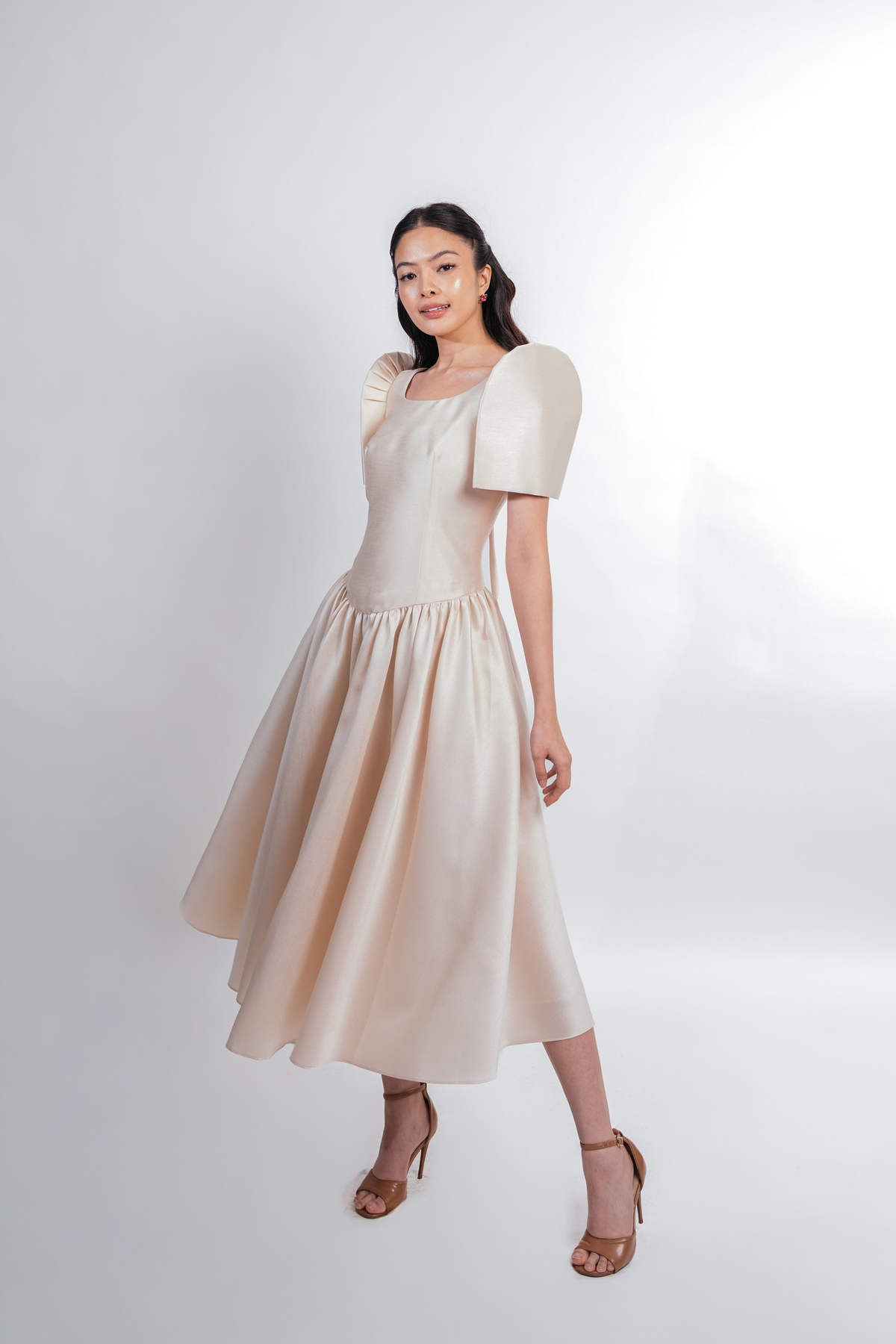 A woman modeling a light beige midi dress with butterfly sleeves, a square neckline, and a gathered skirt, paired with brown heeled sandals against a light gray studio background.