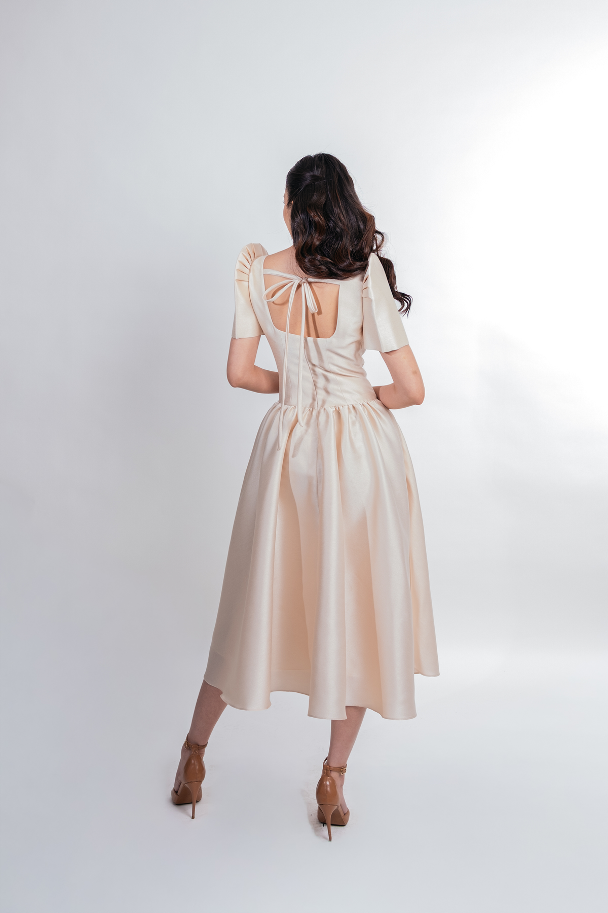 A woman viewed from behind wearing a beige satin midi dress with butterfly sleeves, a tie-back neckline, and a gathered skirt, paired with brown high-heeled sandals against a white studio background.