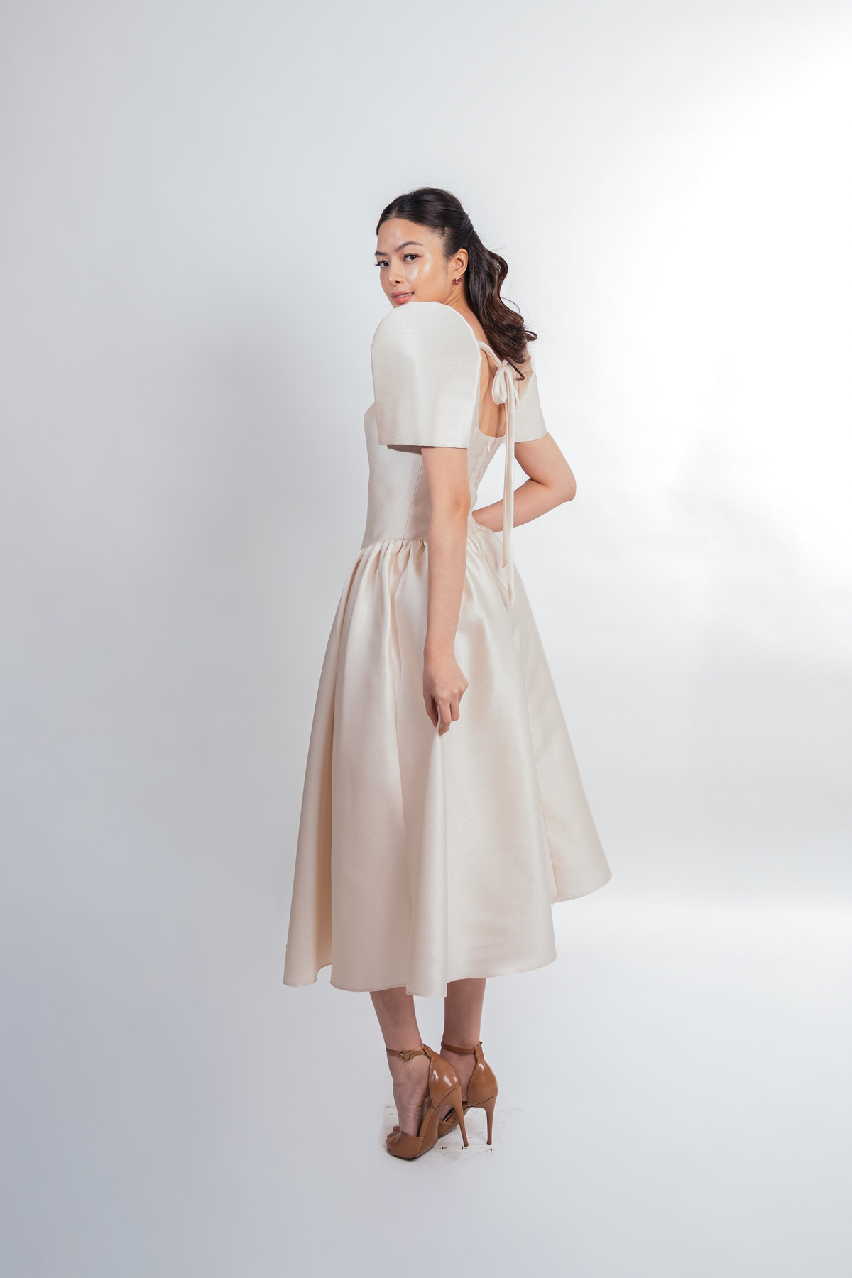 A woman modeling a cream-colored midi dress with short puff sleeves and a gathered skirt, shown from a three-quarter back angle against a light gray studio background.