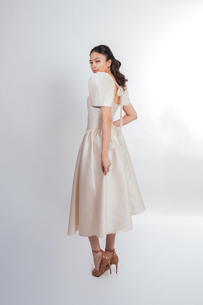 A woman modeling a cream-colored midi dress with short puff sleeves and a gathered skirt, shown from a three-quarter back angle against a light gray studio background.