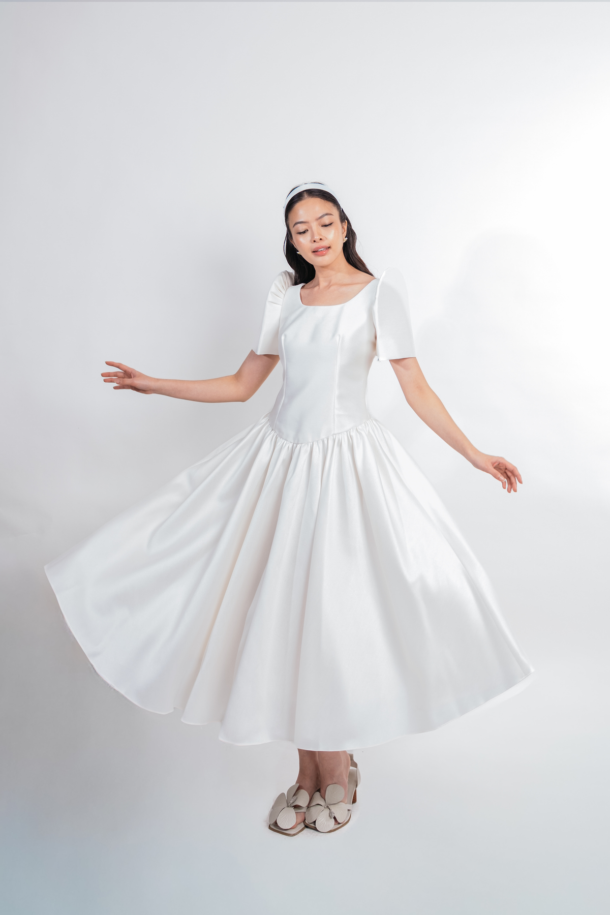 A woman wearing a white short-sleeve midi dress with a structured bodice and a full flared skirt, posed in a twirling motion against a light gray studio background.