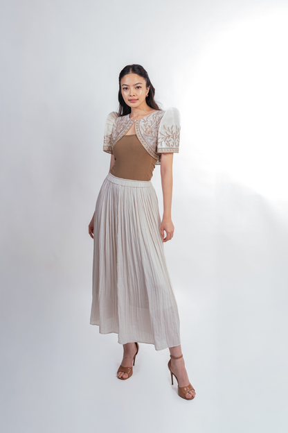 A woman modeling a two-piece outfit consisting of a brown cropped bolero with sheer butterfly sleeves and a matching pleated light beige maxi skirt, paired with brown heeled sandals against a white studio background.