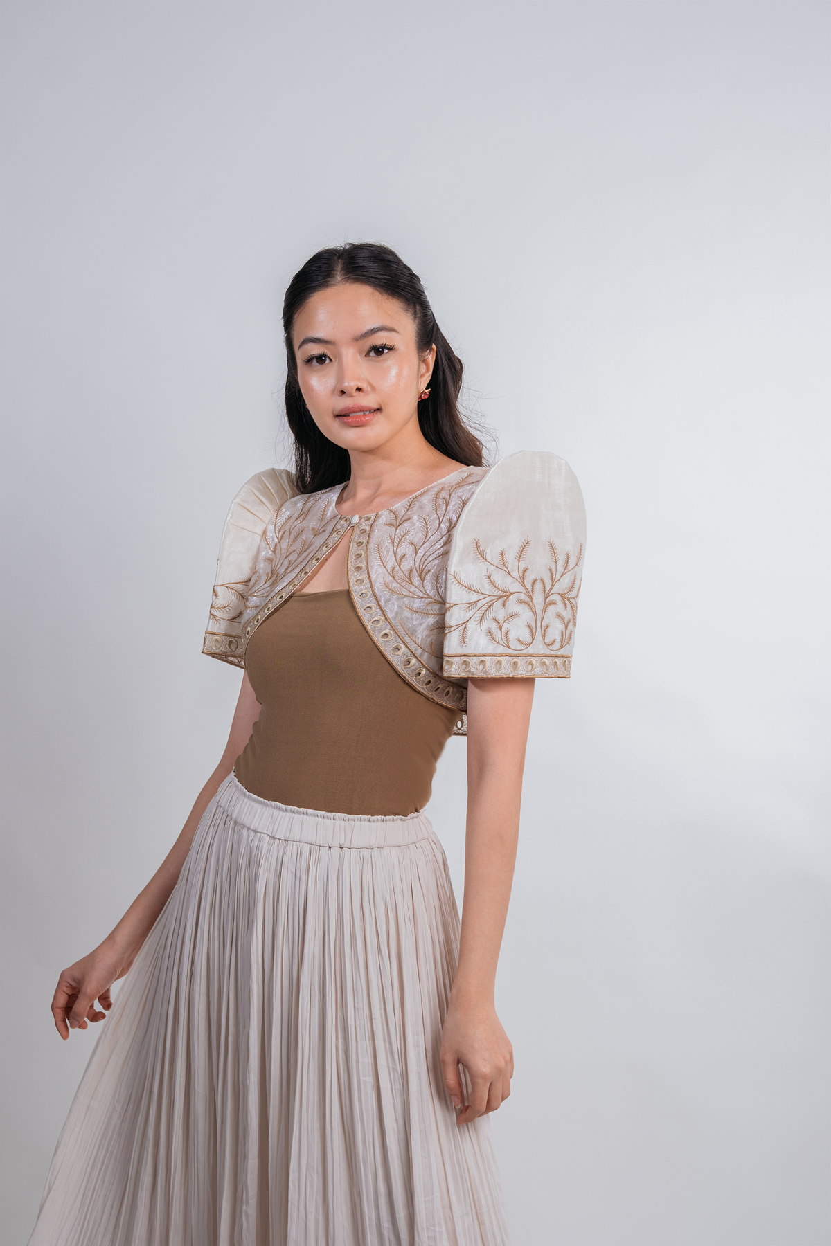 A woman modeling a beige and cream puff-sleeve top with intricate embroidery, paired with a pleated light beige skirt, against a plain light gray studio background.