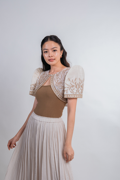 A woman modeling a beige and cream puff-sleeve top with intricate embroidery, paired with a pleated light beige skirt, against a plain light gray studio background.