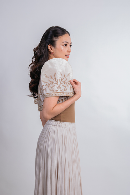A woman in a beige and cream embroidered traditional-style dress, shown from the back and side against a plain light gray studio background.