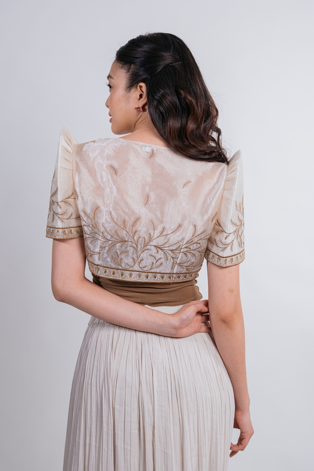 A woman shown from the back wearing a cream and beige embroidered blouse with short puff sleeves and a pleated light beige skirt, against a plain light gray studio background.