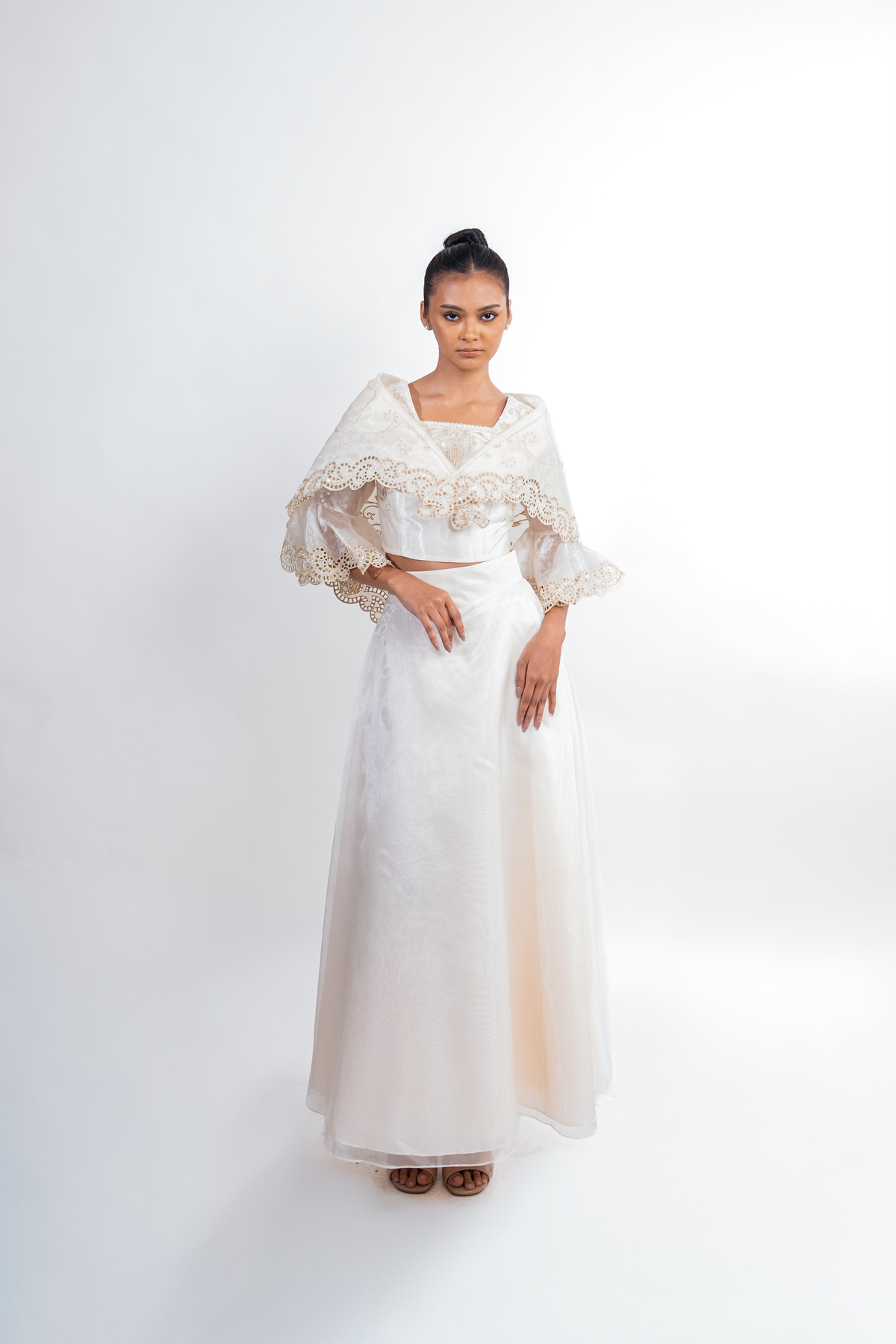 A woman modeling a two-piece white formal dress set with a lace-trimmed crop top and matching flared sleeves, paired with a long flowing skirt, against a plain light gray studio background.