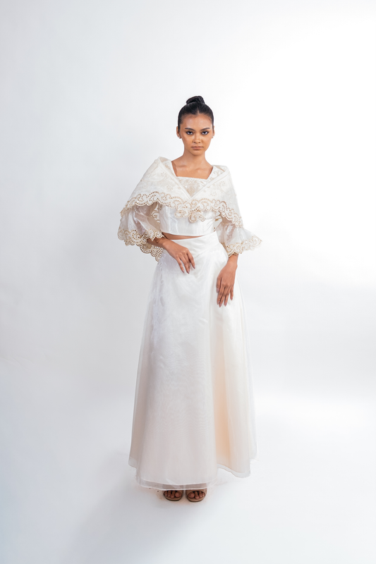 A woman modeling a two-piece white formal dress set with a lace-trimmed crop top and matching flared sleeves, paired with a long flowing skirt, against a plain light gray studio background.