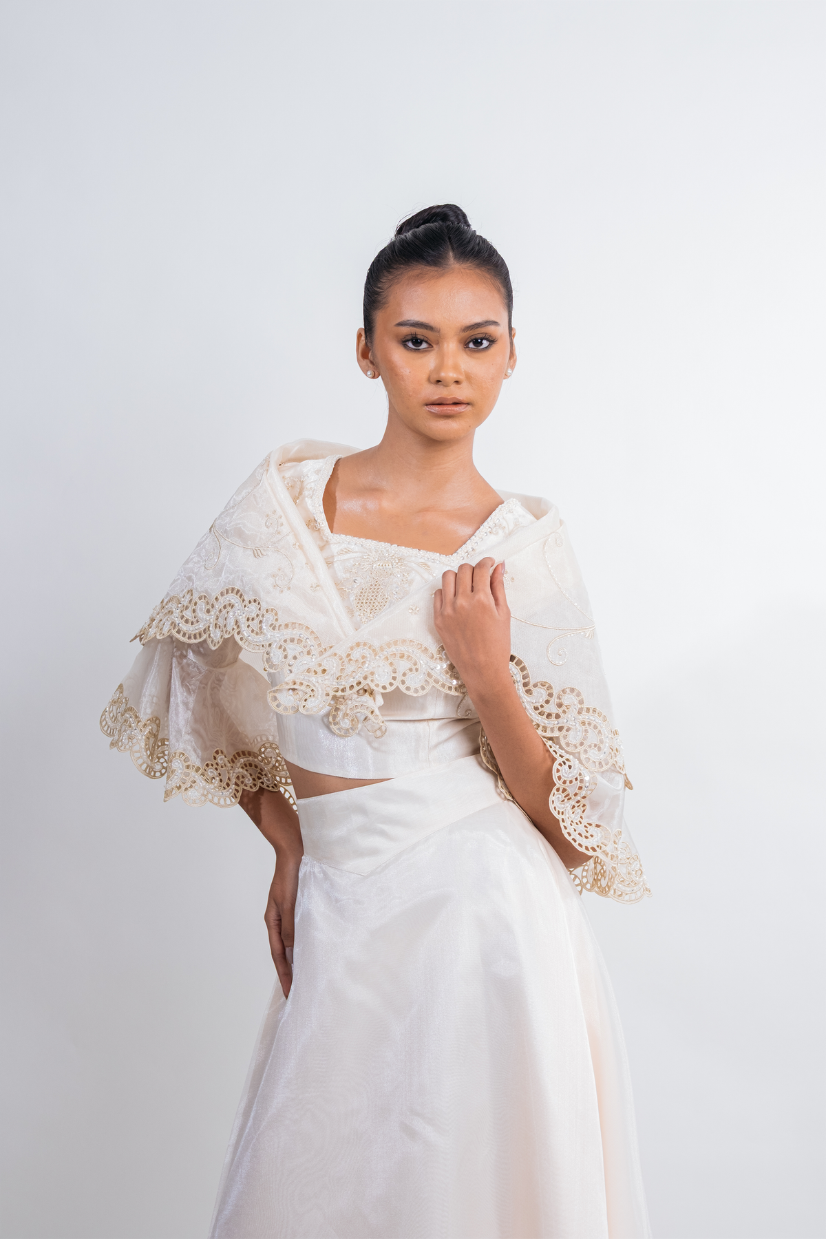 A woman modeling a white two-piece outfit with a lace-trimmed draped capelet and a matching satin skirt, photographed against a plain light gray studio background.