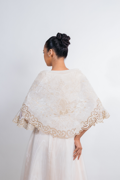 A woman shown from the back wearing a cream lace alampay or shawl over a white dress against a plain light gray studio background.