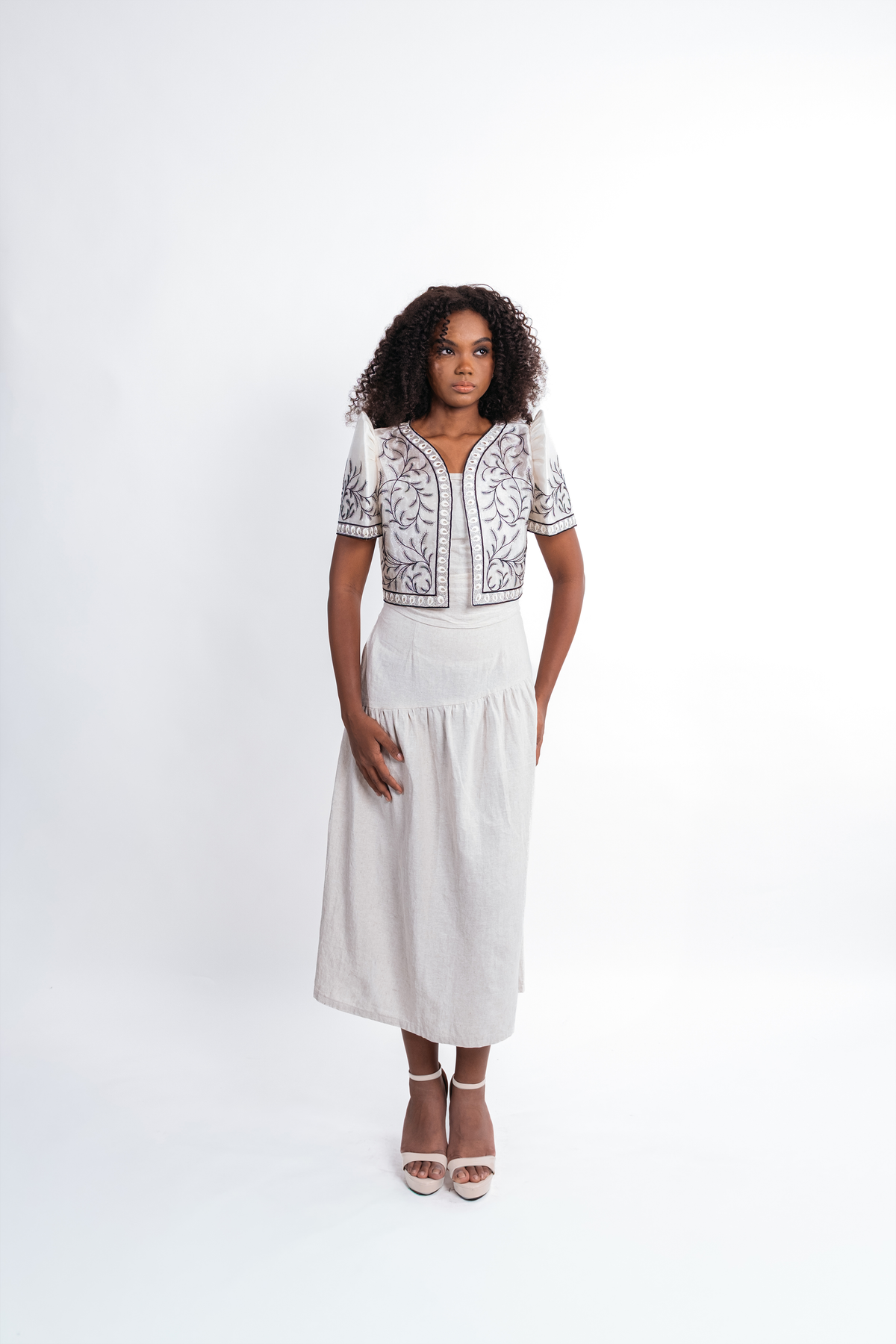 A woman modeling a white midi dress with a bolero top featuring black and gray floral embroidery, paired with white high-heeled sandals, against a plain white studio background.