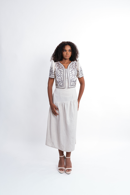 A woman modeling a white midi dress with a bolero top featuring black and gray floral embroidery, paired with white high-heeled sandals, against a plain white studio background.